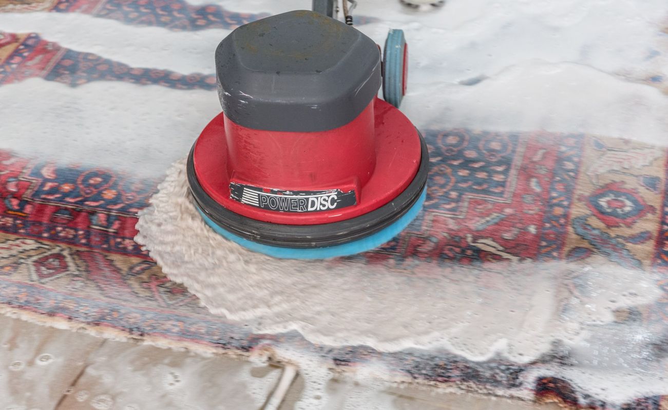 Ein Teppich wird mit einer rotierenden Reinigungsmaschine gereinigt, während Schaum und Wasser auf den Boden spritzen.