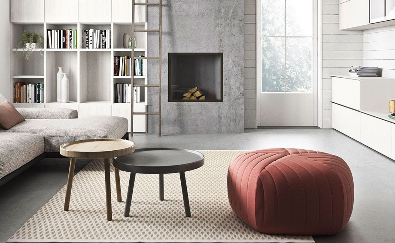 Modernes Wohnzimmer mit einem Sofa, einem runden Tisch, einem Hocker und einem Kamin. Helle Farben und große Fenster sorgen für eine freundliche Atmosphäre.