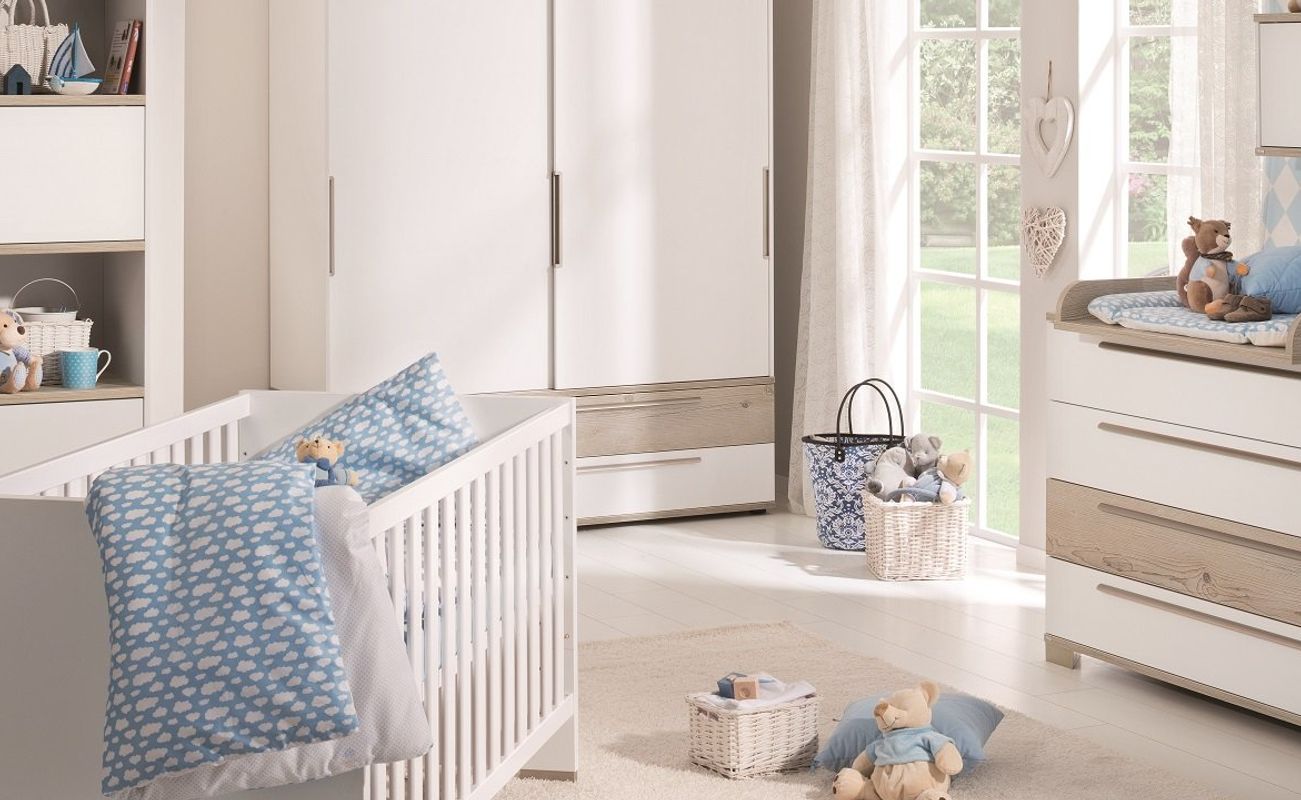 Ein helles Kinderzimmer mit einem weißen Babybett, einer Wickelkommode, einem Regal und einem großen Fenster. Dekor in Blau und Weiß.