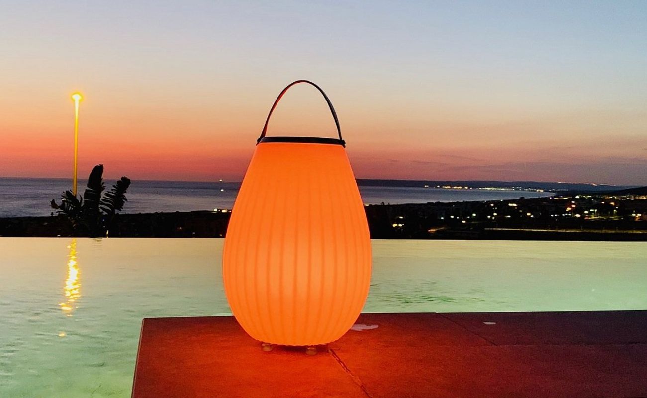 Ein leuchtender, orangefarbener Laternenbehälter steht am Rand eines Infinity-Pools mit Blick auf das Meer und den Sonnenuntergang.