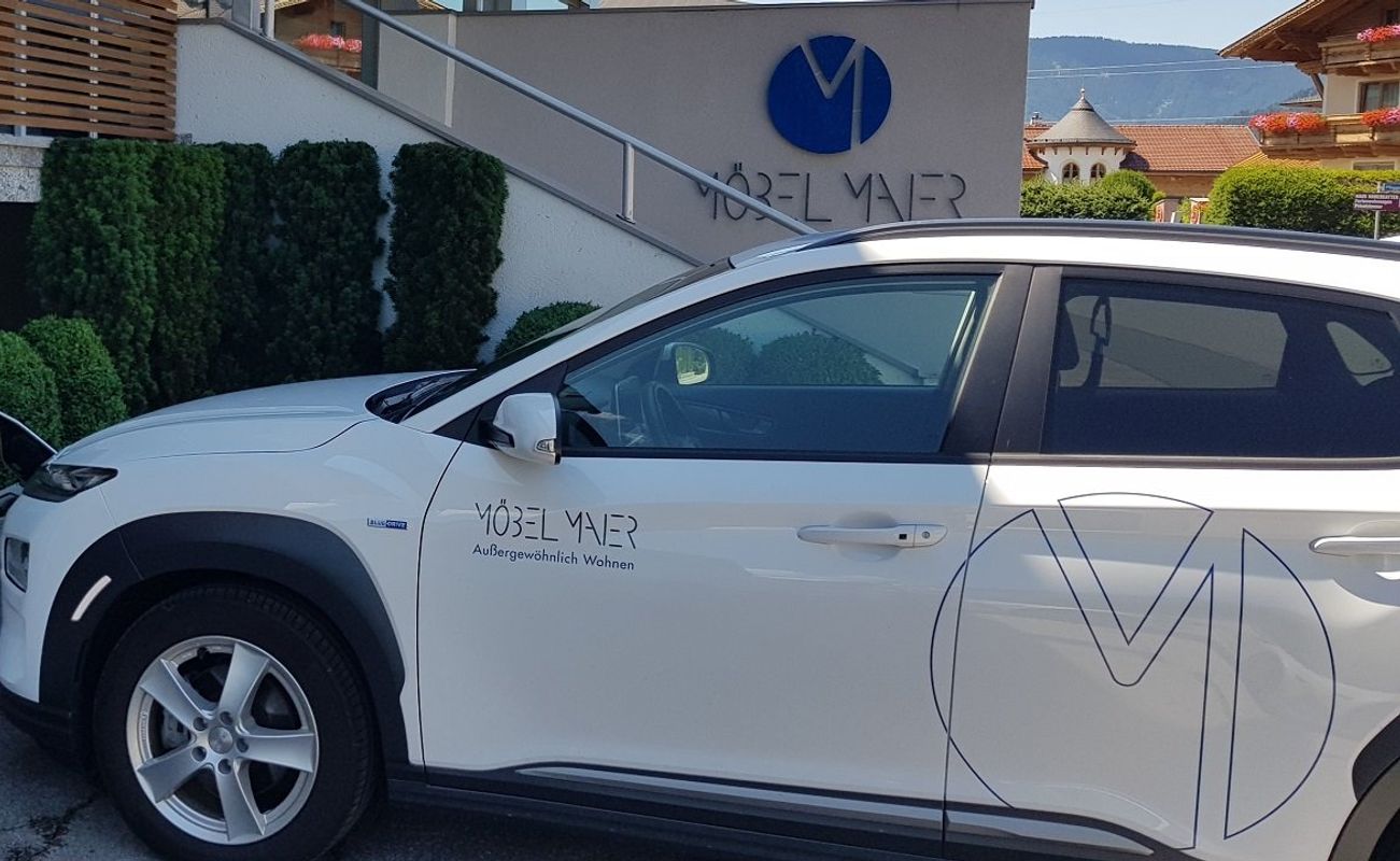 Ein weißes Elektroauto parkt neben einer Ladestation vor einem Gebäude mit dem Schriftzug "Möbel Mayer". Grüne Sträucher im Hintergrund.